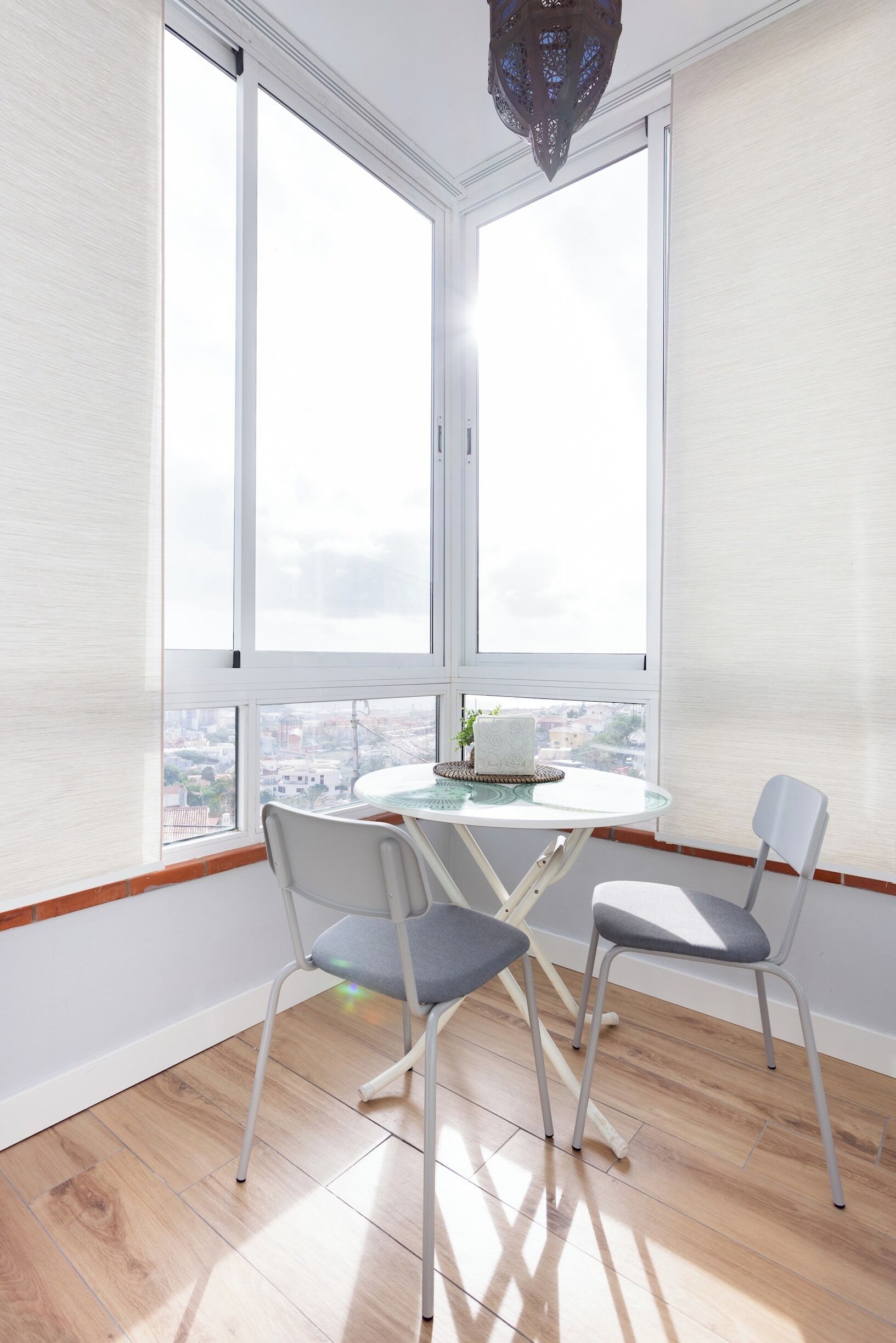 Quarto com mesa redonda e duas cadeiras, grandes janelas com vista para a cidade e luminária suspensa.