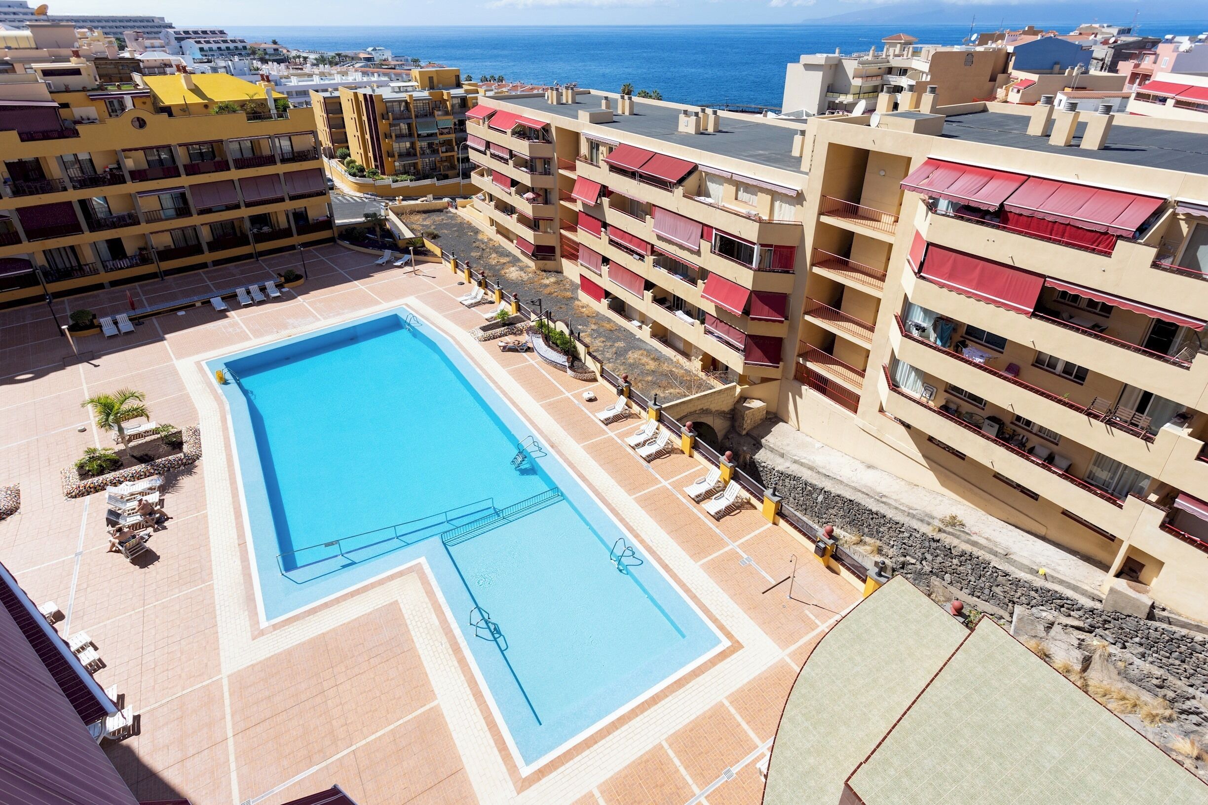 A rooftop swimming pool surrounded by multi-story buildings with balconies, a clear view of the sea, and a paved area with lounge chairs.