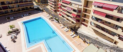 A rooftop swimming pool surrounded by multi-story buildings with balconies, a clear view of the sea, and a paved area with lounge chairs.