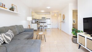 Ein modernes Wohnzimmer mit einer grauen Couch, einem kleinen runden Tisch und einem Fernseher auf einem Schrank. Im Hintergrund ist eine Küche mit weißen Schränken und einem Essbereich zu sehen.