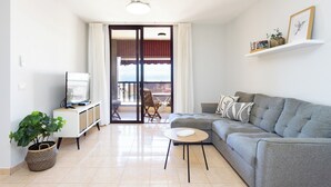 Ein modernes Wohnzimmer mit einem grauen Sofa, einem kleinen runden Tisch, einem Fernseher auf einem weißen Schrank, einer Topfpflanze und einem Balkon mit Aussicht.
