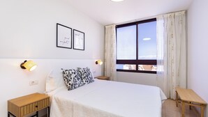 Ein Schlafzimmer mit einem Bett, zwei Nachttischen, einer Wandleuchte, einem Fenster mit Vorhängen und zwei gerahmten Bildern an der Wand.