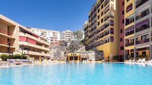 Ein Schwimmbecken, umgeben von mehrstöckigen Gebäuden mit Balkonen, ein felsiger Hügel im Hintergrund und ein klarer, blauer Himmel.