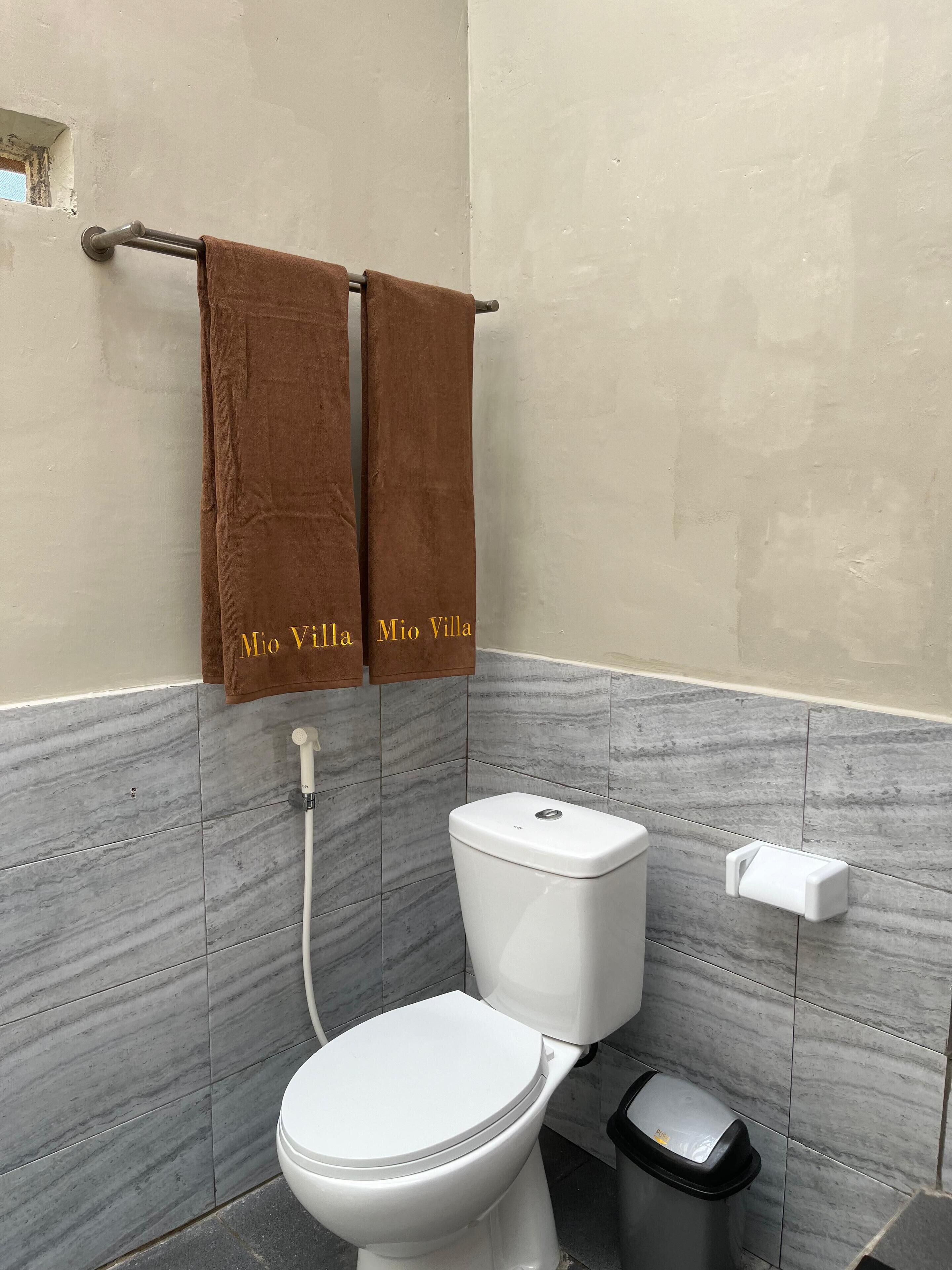 Une salle de bain équipée de toilettes, d’un bidet et d’une poubelle. Deux serviettes sont suspendues à un porte-serviettes, et l’une d’elles porte l’inscription « Mio Villa ». Les murs sont recouverts de carreaux imitant le marbre.