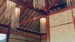A rustic interior with a high wooden ceiling, woven ceiling panels, and hanging woven light fixtures. There are wooden tables and chairs arranged for seating. - Mio Villa Tomang Omang Lombok (Selong Belanak)