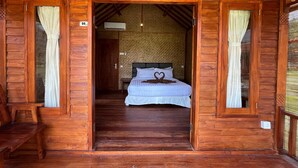A wooden cabin with a bed in the center, a bench to the left, and a door leading to another room. - Mio Villa Tomang Omang Lombok (Selong Belanak)