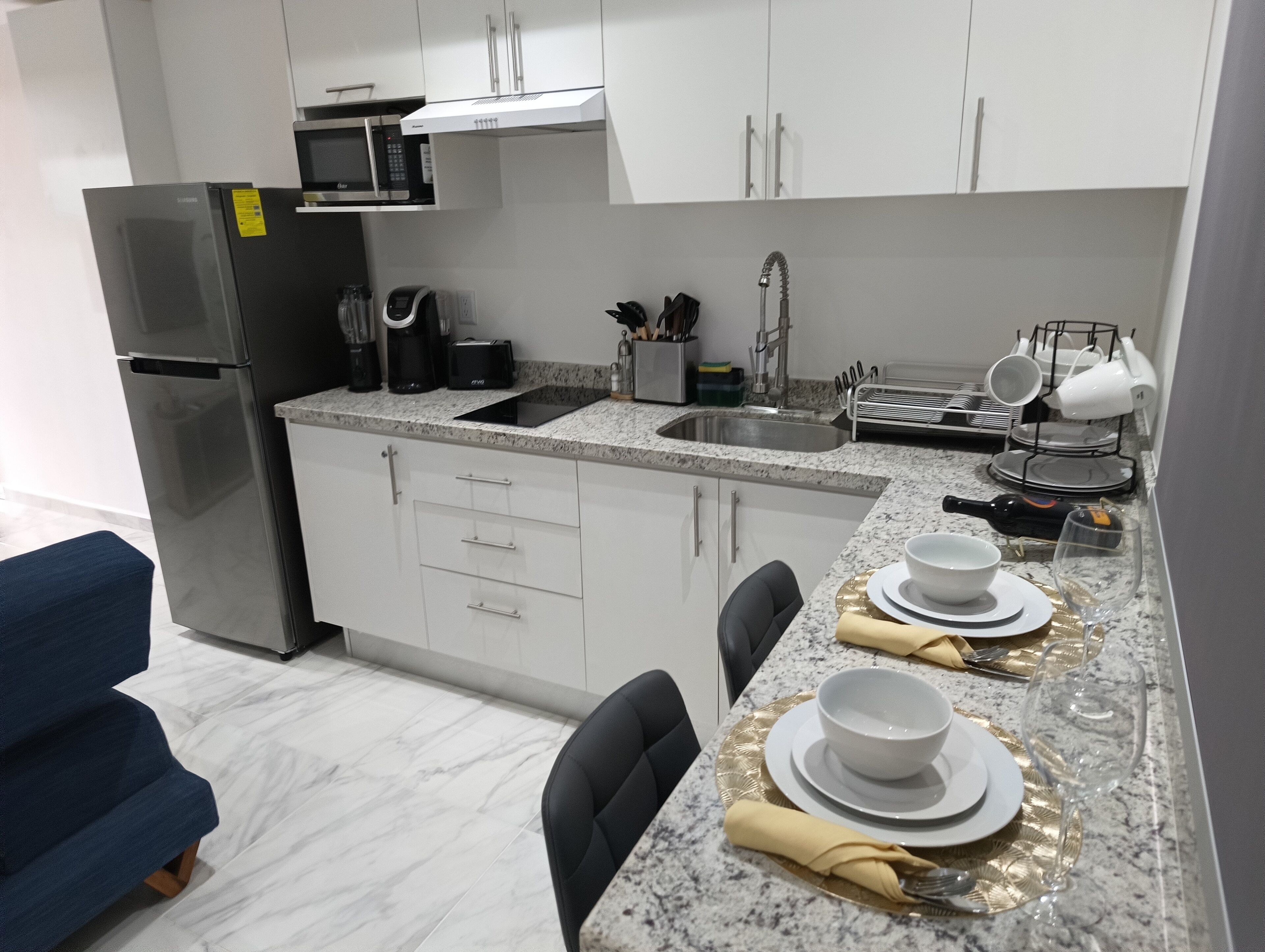 Una cocina moderna con gabinetes blancos, un refrigerador de acero inoxidable, un microondas y un fregadero. Hay una zona de comedor con una mesa preparada para dos personas, incluyendo platos, tazones y una copa de vino.