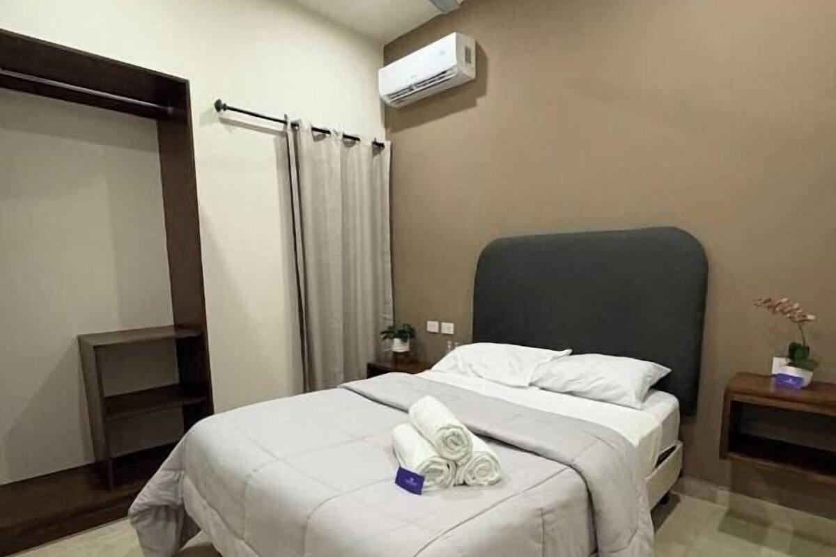 A hotel room with a bed, a ceiling fan, an air conditioning unit, a small bedside table with a plant, and a wardrobe.