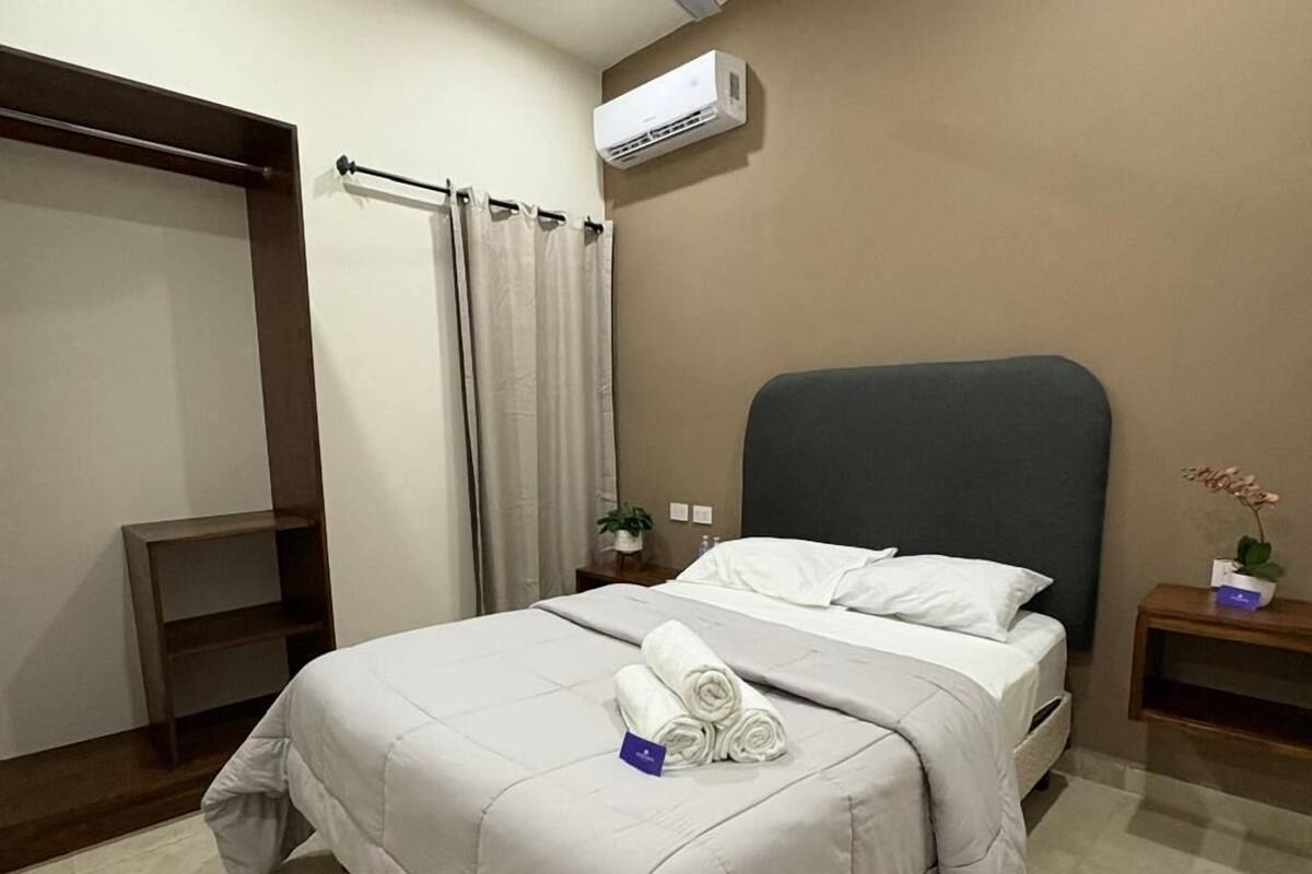 A neatly made bed with a white comforter and rolled towels, a ceiling fan, an air conditioning unit, a small bedside table with a plant, and a dark headboard.