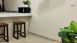 A modern kitchen with a flat-screen TV mounted on the wall, a microwave, and a sink. There are two wooden stools and a white table with an hourglass and a potted plant. - Hortensias Hotel and Suites (Tapachula)
