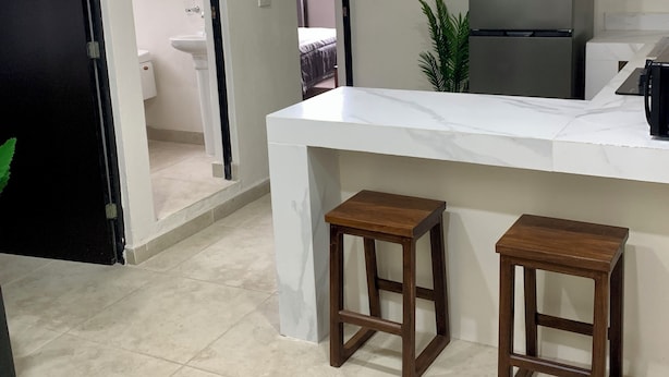 A kitchen with a white countertop, two wooden stools, a refrigerator, and a potted plant. - Hortensias Hotel and Suites (Tapachula)
