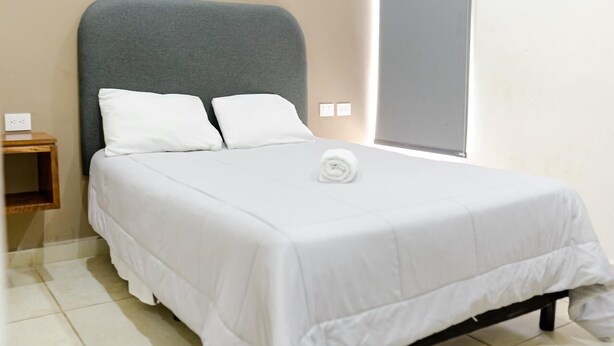 A neatly made bed with white bedding and pillows, a grey headboard, and a small wooden nightstand with a power outlet. - Hortensias Hotel and Suites (Tapachula)