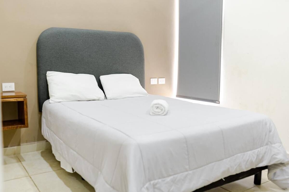 A neatly made bed with white bedding and pillows, a grey headboard, and a small wooden nightstand with a power outlet.
