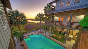 A pool with a view of the sunset, a deck with lounge chairs, and a building with a balcony. - Fish Camp by Avantstay Unique Escape, Pool + Views! (Port Aransas)