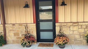 Eine Haustür mit einer satinierten Glasscheibe, zwei Blumentöpfe mit Blumen, eine Fußmatte mit Schriftzug und eine Steinmauer.