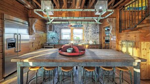 A rustic kitchen with a central island, bar stools, a stainless steel refrigerator, and a stone backsplash. - Fish Camp Buyout by Avantstay Unique 6bdr Escape, Private Pool + Views! (Port Aransas)