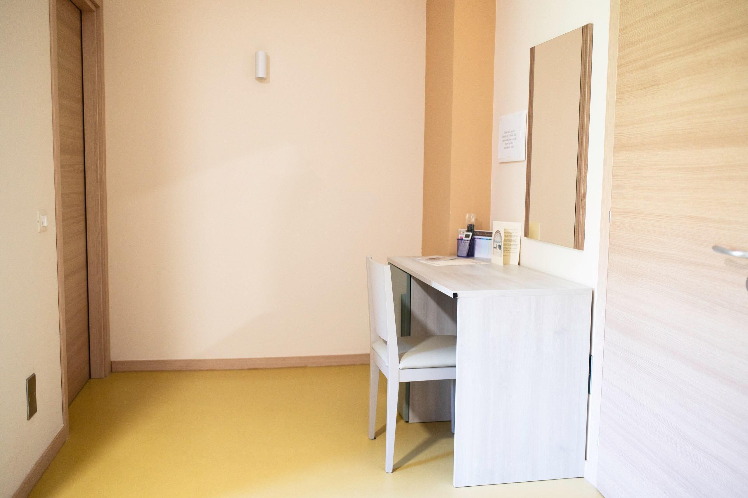 A small, compact room with a wooden door, a white desk with a chair, and a mirror on the wall.