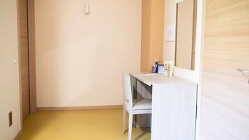 A small, compact room with a wooden door, a white desk with a chair, and a mirror on the wall.