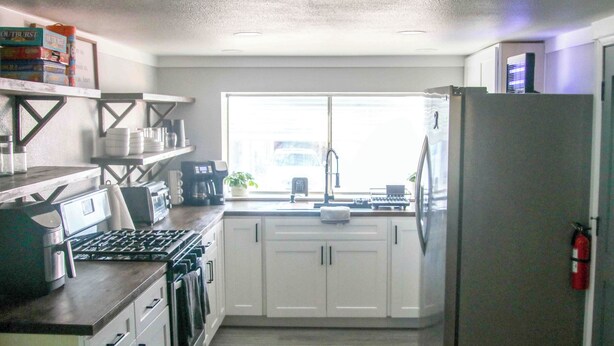 A kitchen with white cabinets, a stainless steel refrigerator, a gas stove, and a window providing natural light. - Welcome to The Cozy Cove, Your Lakeside Retreat (Havasu Lake)