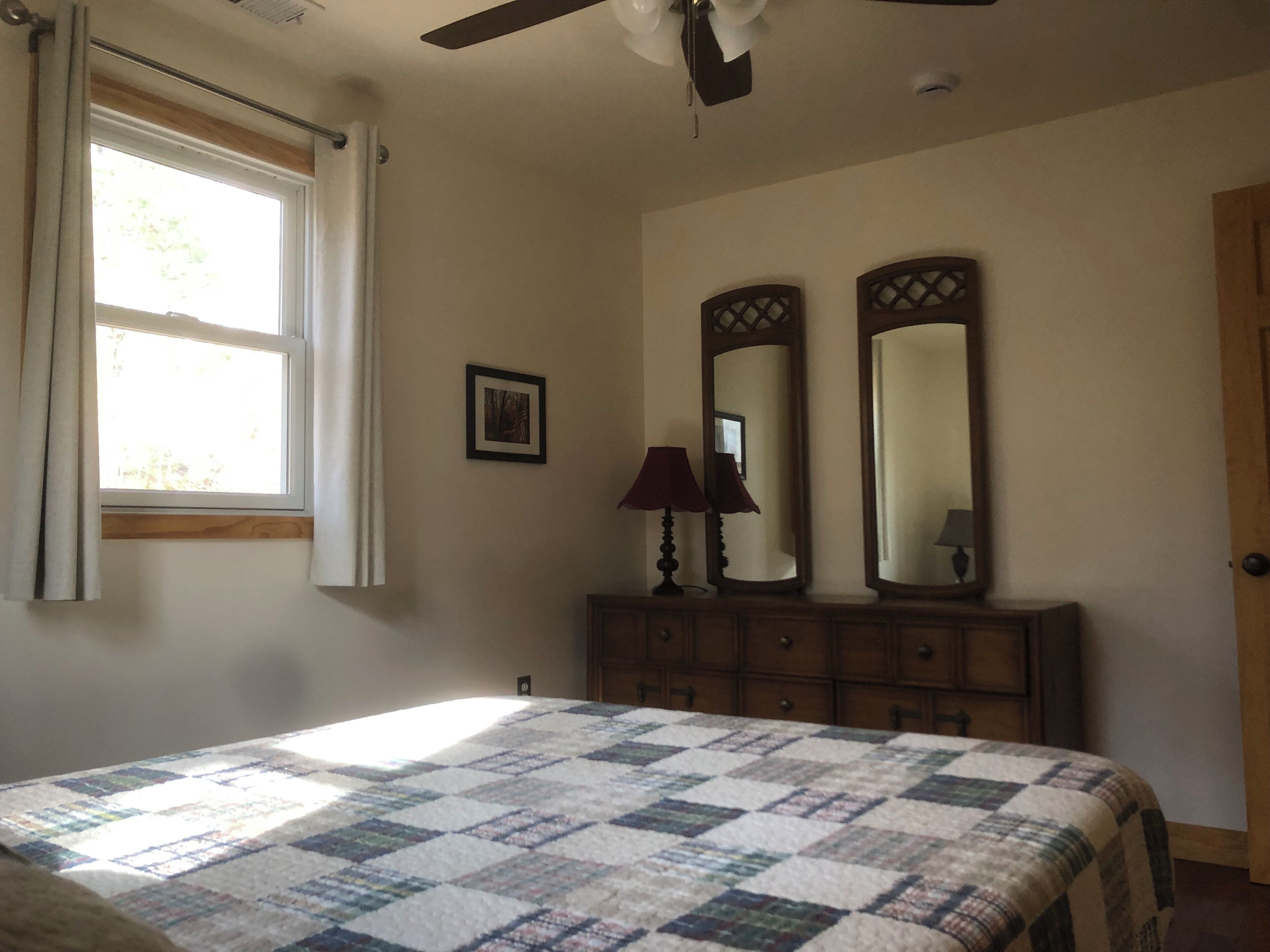 A bedroom with a bed, a window with curtains, a dresser, two mirrors, and a ceiling fan.