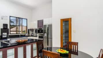 A modern kitchen with dark wooden cabinets, a black countertop, and a dining table with chairs. There is a fruit bowl on the table.