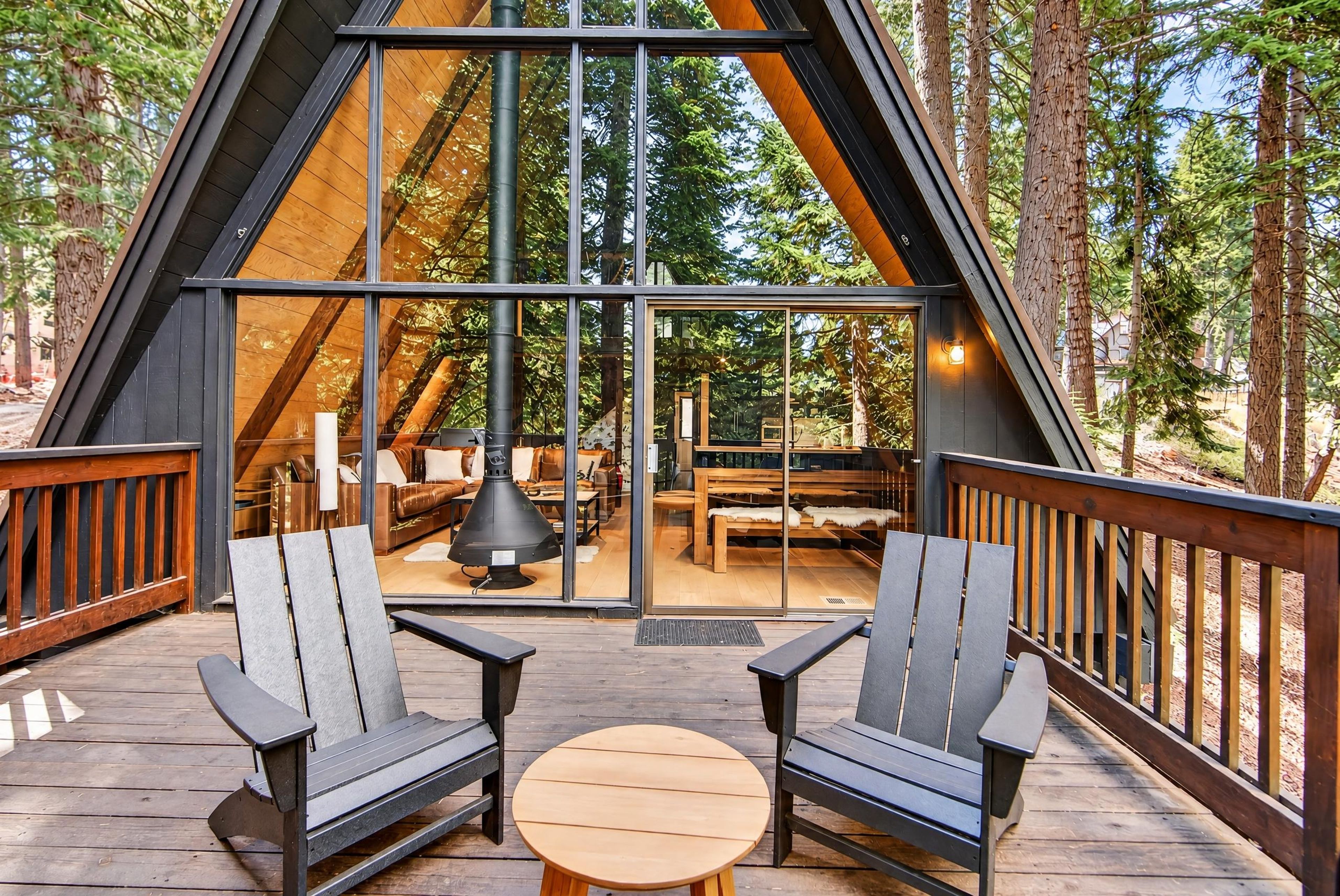Une terrasse en bois avec deux chaises et une table, entourée de murs de verre donnant sur un espace intérieur aménagé avec des sièges et une cheminée.