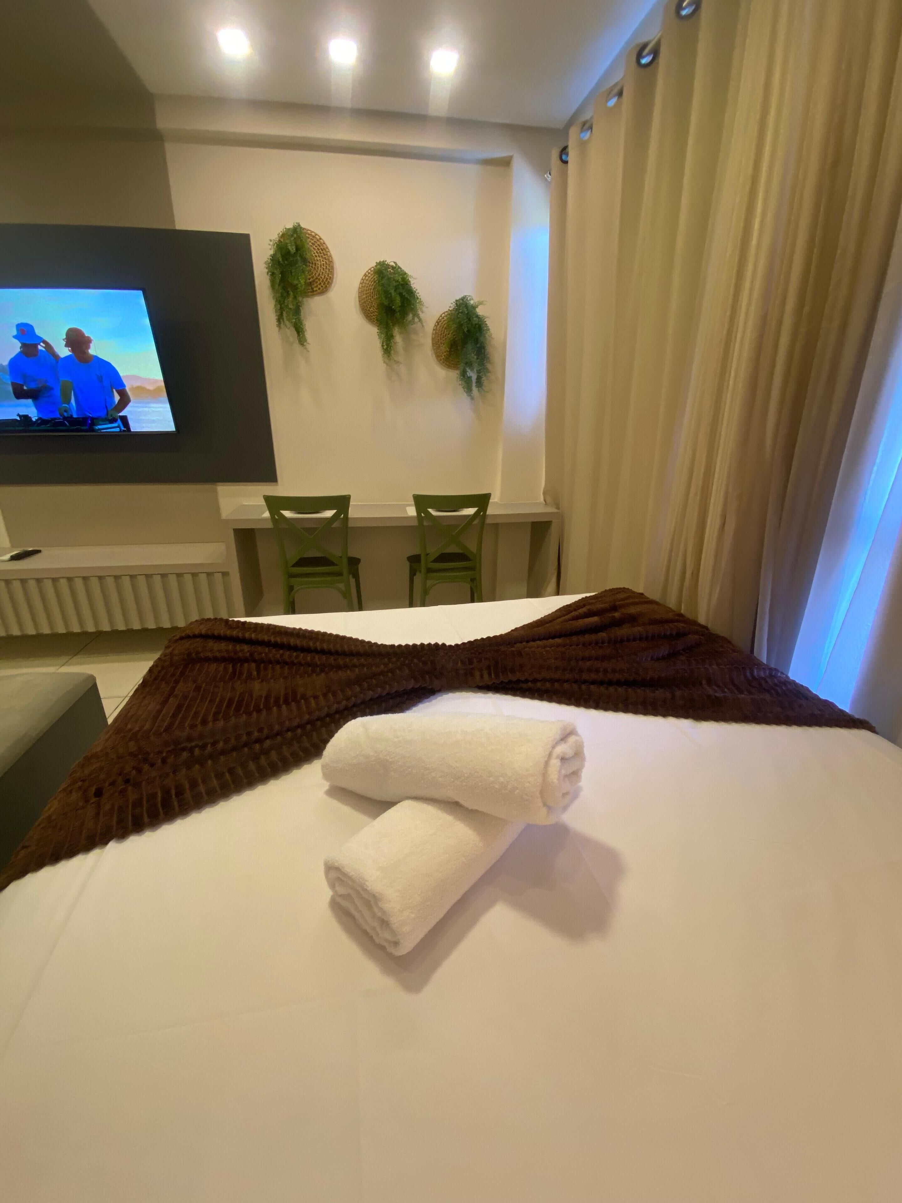 A hotel room with a bed, a TV mounted on the wall, two chairs, and a window with curtains.