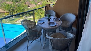 A balcony with wicker chairs and a table set for dining, overlooking a pool and a tropical landscape with palm trees. - Flat 211 Vista Mar EcoResort by La Playa (Tamandaré)