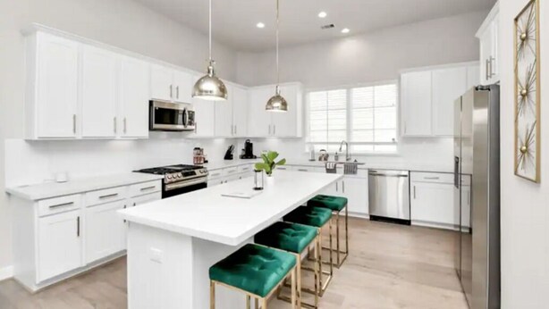 A modern kitchen with white cabinets, a central island with green bar stools, stainless steel appliances, pendant lighting, and a window providing natural light. - The Houston Hangout: Modern Comfort & Style (Houston)