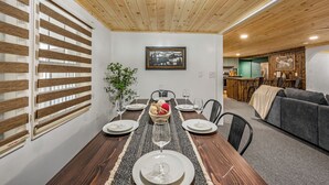 A dining area with a wooden ceiling, a large window with blinds, a framed picture on the wall, and a dining table set with plates, glasses, and a centerpiece. - Location! ★ Game R. ★ Movie R. ★ Hot Tub ★ Fire Pit ★ King Beds ★ Dollywood! (Sevierville)