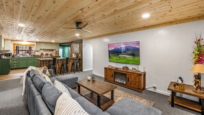 A modern living room with a wooden ceiling, a large flat-screen TV, a fireplace, and a kitchen area in the background. - Location! ★ Game R. ★ Movie R. ★ Hot Tub ★ Fire Pit ★ King Beds ★ Dollywood! (Sevierville)