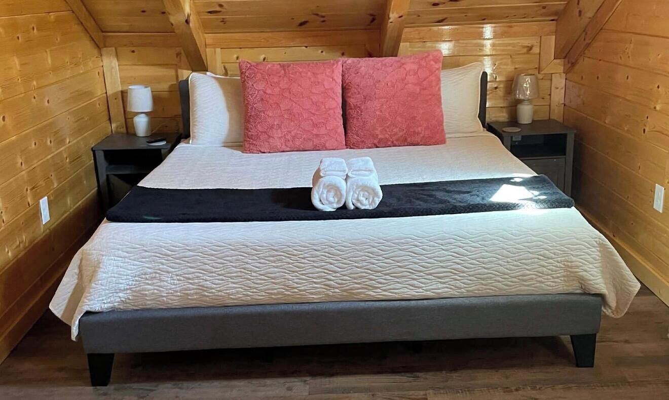 Une chambre dans une cabane en bois, avec un lit, des oreillers, une table de chevet et une lampe.