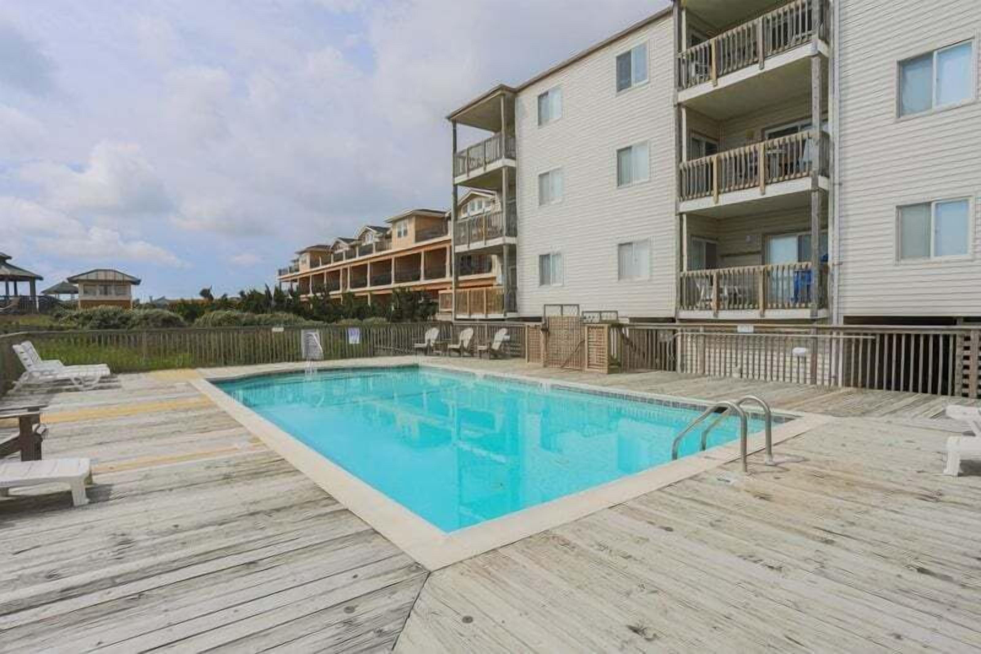 A swimming pool with a wooden deck, lounge chairs, and a multi-story building with balconies.