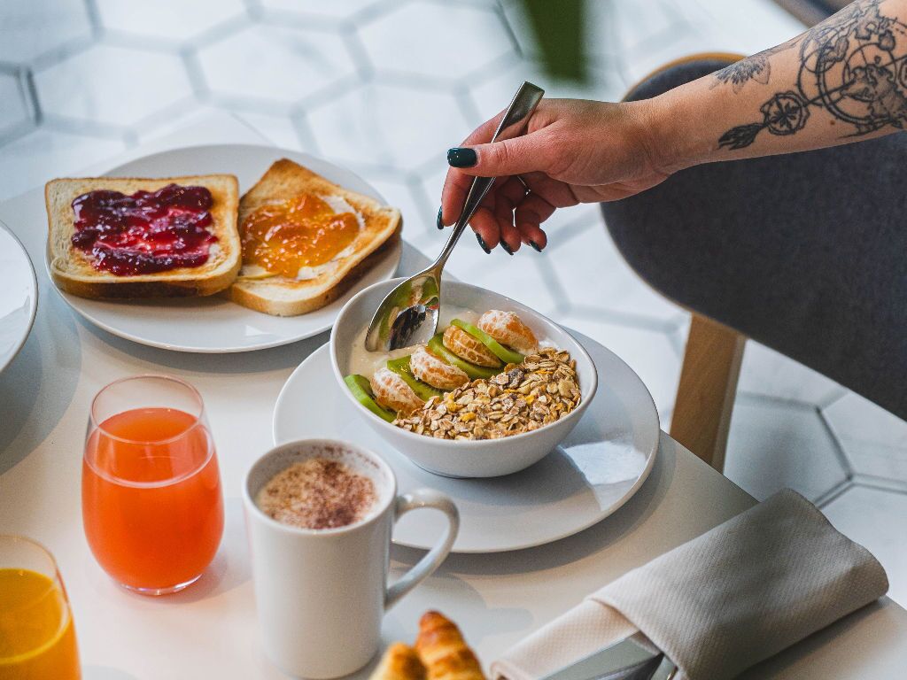 Desayuno con tostadas, mermelada, cereal con fruta, un vaso de jugo de naranja y una taza de café.