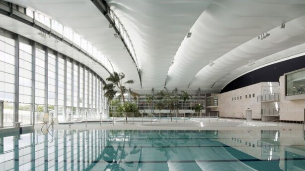 An indoor swimming pool with a curved ceiling, large windows, and palm trees.