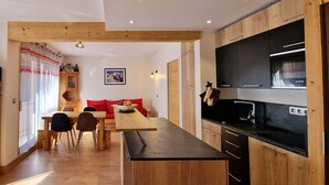 A modern kitchen with wooden cabinets, a black countertop, and a built-in oven. There is a dining area with a wooden table and chairs, and a red sofa.