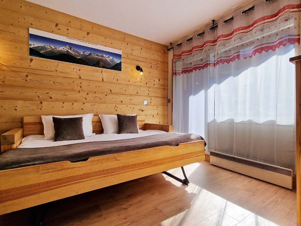 A wooden bed with a headboard, a mounted picture of a mountain landscape, and a window with sheer curtains.