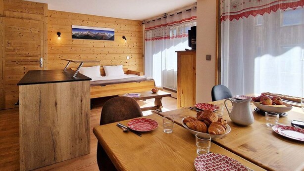 A cozy room with a bed, wooden walls, a dining table with plates and a pitcher, and a window with curtains.