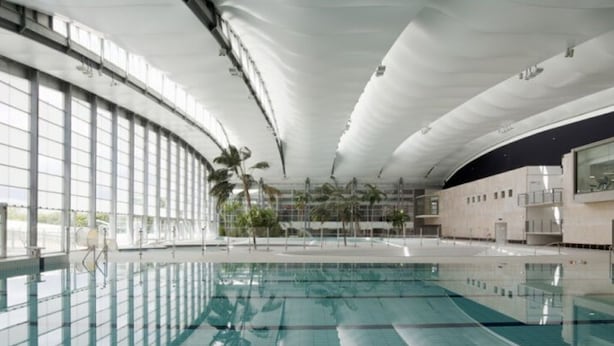 An indoor swimming pool with a curved ceiling, large windows, and palm trees.