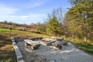 A fire pit with stone seating surrounded by gravel, set in a natural landscape with trees and grass. - Byers Haven | Hocking Hills, 5BR, Hot Tub, Firepit (Logan)