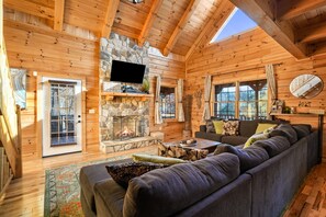 A log cabin interior with a stone fireplace, a large sofa, and a television mounted on the wall. - Byers Haven | Hocking Hills, 5BR, Hot Tub, Firepit (Logan)