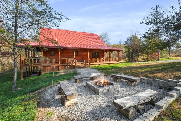 A log cabin with a red roof, a deck, and a fire pit area with benches.