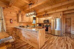 A spacious kitchen with wooden cabinetry, a central island, stainless steel appliances, and a hanging light fixture. - Byers Haven | Hocking Hills, 5BR, Hot Tub, Firepit (Logan)