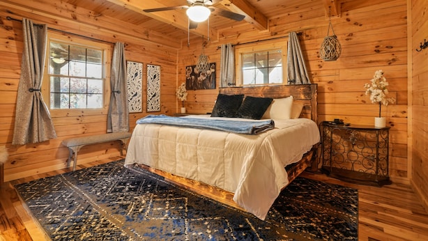 Een slaapkamer in een houten hutje met een bed, een plafondventilator en een raam met gordijnen.