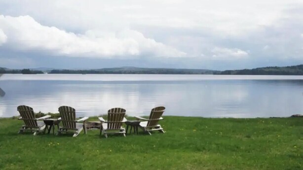 Une vue sur le lac, avec quatre chaises Adirondack et une petite table, le tout entouré d’une prairie et surplombant un lac paisible sous un ciel nuageux.