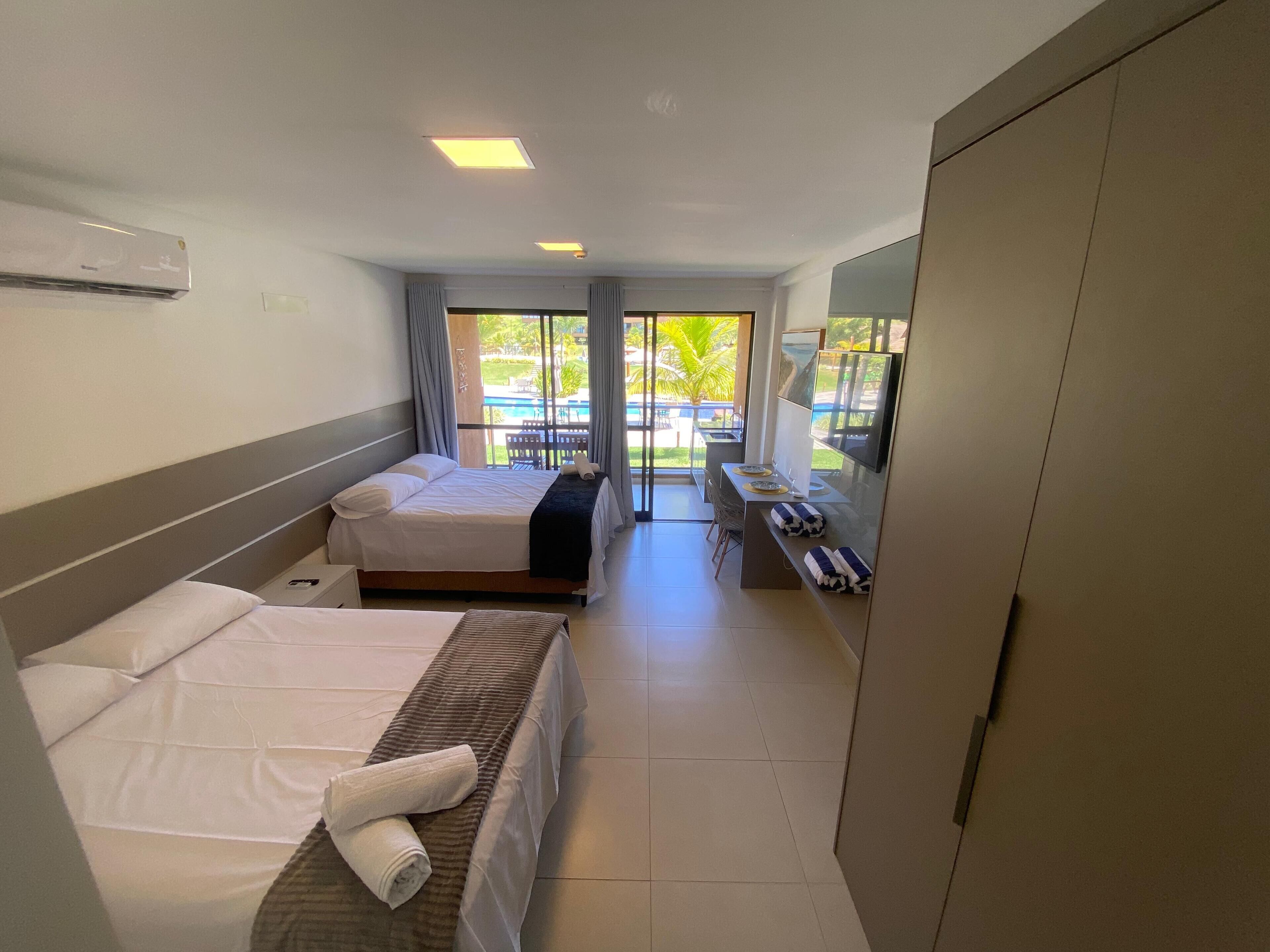 A hotel room with two beds, a desk with a TV, and a view of a pool and palm trees.