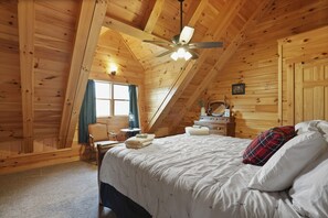 Een knus slaapkamer onder dak met een schuine wand, houten lambrisering, een bed met wit beddengoed, een plafondventilator en een kleine tafel met stoel.