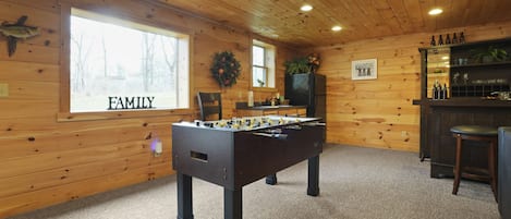 A cozy room with wooden walls, a foosball table, a bar area, and a window with a 'FAMILY' sign.