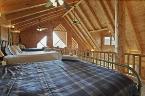 A high-ceilinged wooden cabin bedroom with a large bed, a smaller bed, and a view of the outdoors through a window.