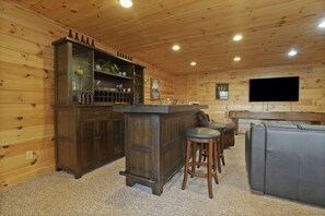 A wooden-paneled room with a bar area, stools, and a flat-screen TV mounted on the wall.